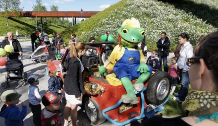 Auf dem Foto ist Buga-Maskottchen Froschkönig Fritz auf einem Auto sitzend zu sehen - umringt von Kindern und Eltern.