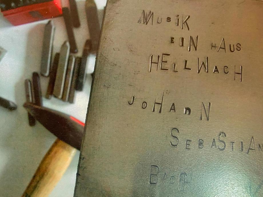 Musik - ein Haus hellwach Auf einer Metalltafel ist ein Text in Schlaggravur eingearbeitet: Musik - ein Haus hellwach - Johann Sebastian Bach. Im Hintergrund sind unscharf die Schlagwerkzeuge zu erkennen.