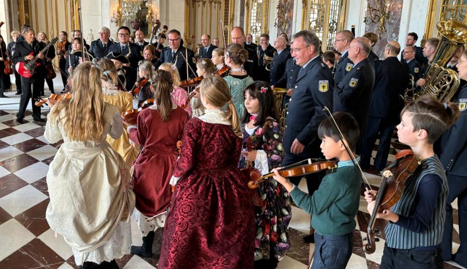 Mitglieder der Jugendnachwuchsorchester in historischen Gewändern strömen in einen prächtigen Saal zu uniformierten Mitgliedern des Bundespolizieorchesters