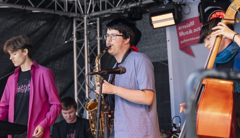 Auf einer bunten Konzertbühne steht wird zwischen anderen Bandenmitgliedern ein Saxophonsolo von einem jungen Mann gespielt.