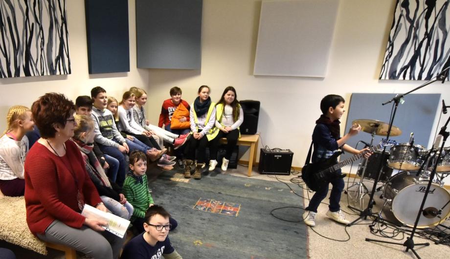 Eine Grundschulklasse ist zu Besuch in einem Tonstudio. Ein sehr junger E-Gitarrist ist dabei in voller musikalischer Aktion.