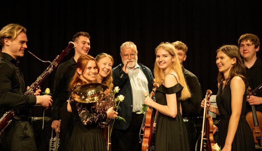 Einige junge klassische Musikerinnen und Musiker freuen sich über einen gelungenen Auftritt und posieren mit ihren Instrumenten. In ihrer Mitte der Leiter der Gruppe aus Opole namens Hubert Prochota. Er ist weißhaarig und hat einen weissen Bart. Auch er freut sich sichtlich!