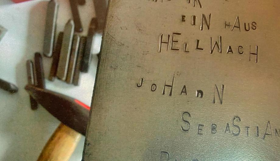 Auf einer Metalltafel ist ein Text in Schlaggravur eingearbeitet: Musik - ein Haus hellwach - Johann Sebastian Bach. Im Hintergrund sind unscharf die Schlagwerkzeuge zu erkennen.