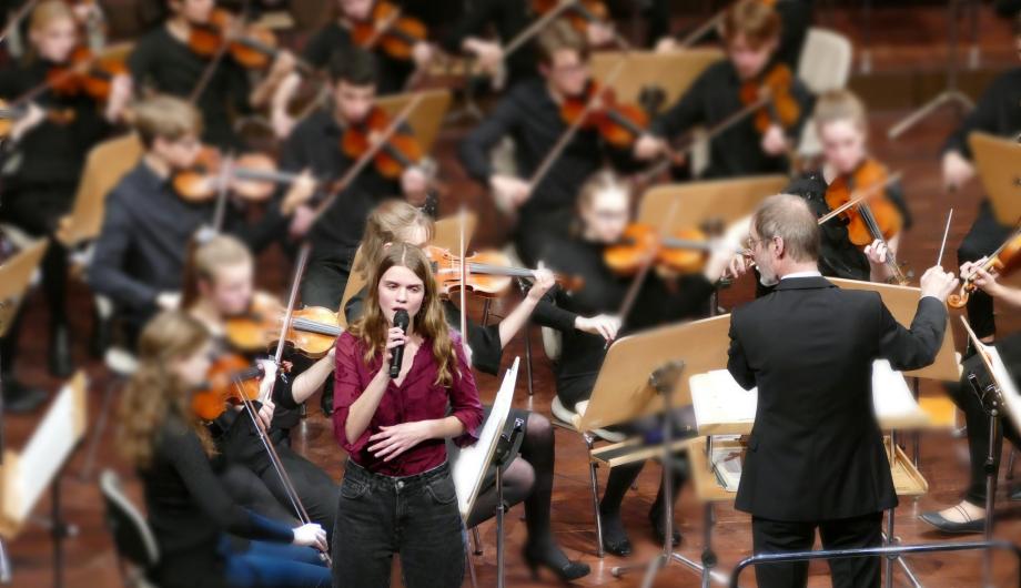 Eine junge Sängerin singt vor einem großen Orchester - der Dirigent schaut zu Ihr