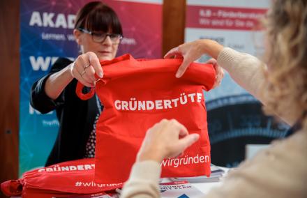 Eine Frau überreicht einer Person eine rote Tasche mit dem Aufdruck 'Gründertüte'
