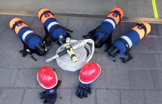 Vier neue Atemschutzgeräte für die Jugendfeuerwehr liegen auf dem Boden, danaben rote Helme, Handschuhe und ein Feuerwehrschlauch mit Spritze.