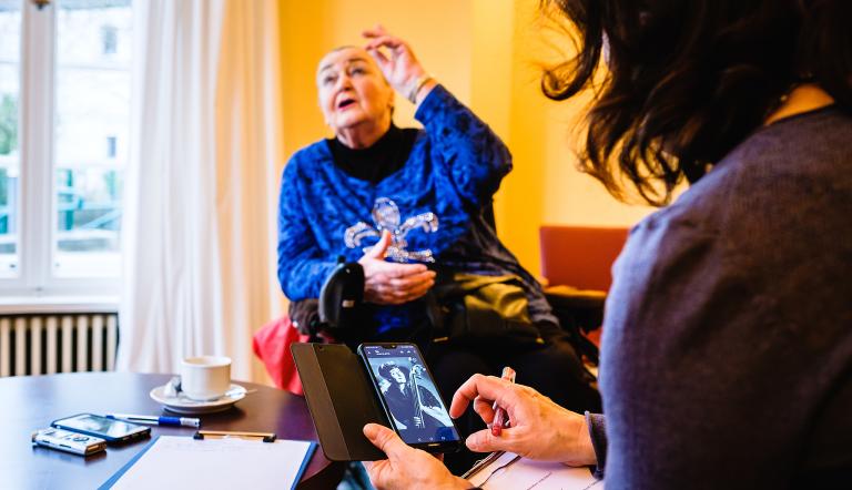 Das Bild zeigt eine Frau, die auf einem Handy im Internet surft, sowie eine ältere sitzende Dame, die etwas erzählt.