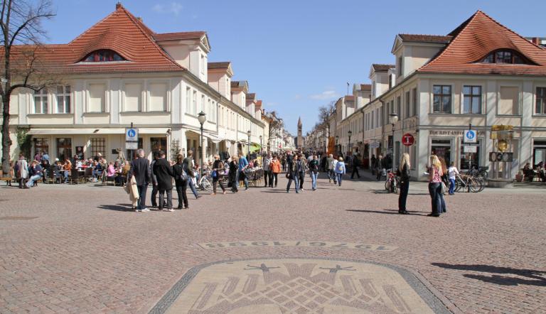 Das Farbfoto zeigt die Brandenburger Straße, von kleinen Geschäfts-und Wohnhäusern gesäumt mit einigen Gästen bei bestem Wetter.