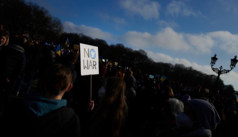 No War - das Schild einer Demonstrantin leuchtet aus einer Menge Demonstrierender. Eine Friedenstaube ist in blau in das "O" von No War gemalt.